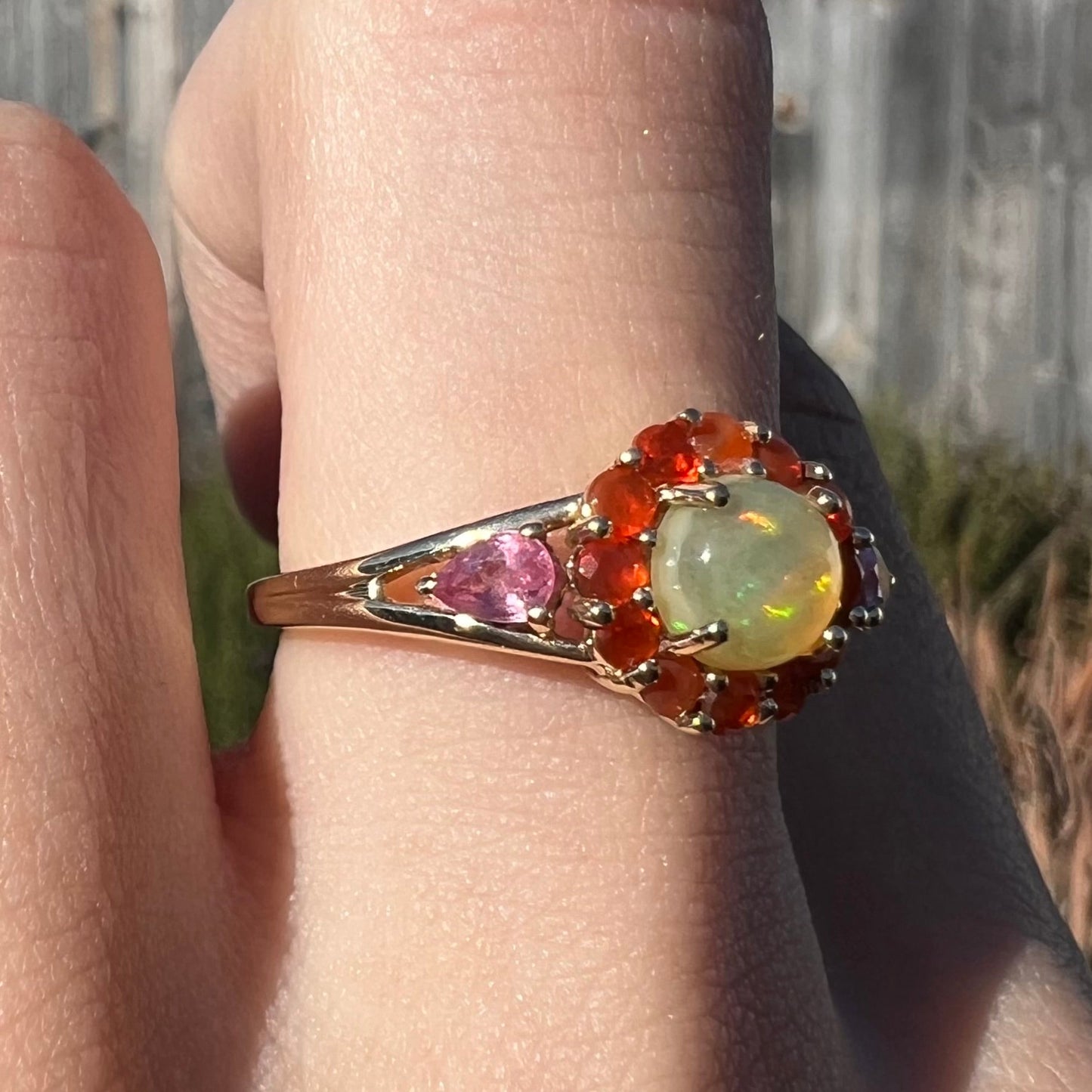 A yellow gold ring mounted with a round Ethiopian opal in a halo of faceted Mexican fire opals. Two pear shaped pink tourmalines accent the sides of the ring.