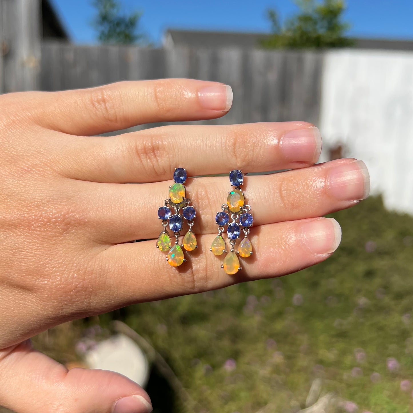 A pair of dangling sterling silver Ethiopian opal and tanzanite chandelier earrings in sunlight.