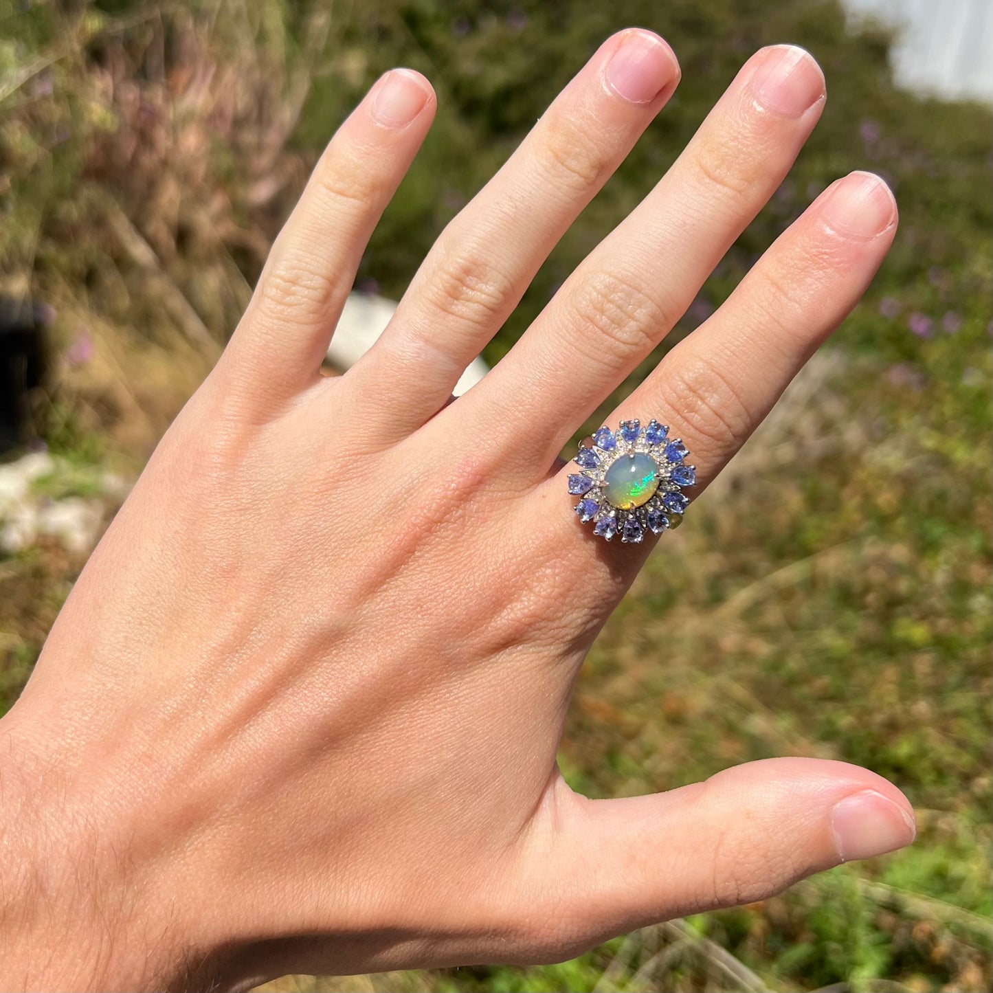 An Ethiopian opal and tanzanite starburst ring, worn on a hand at a distance in the sunlight.