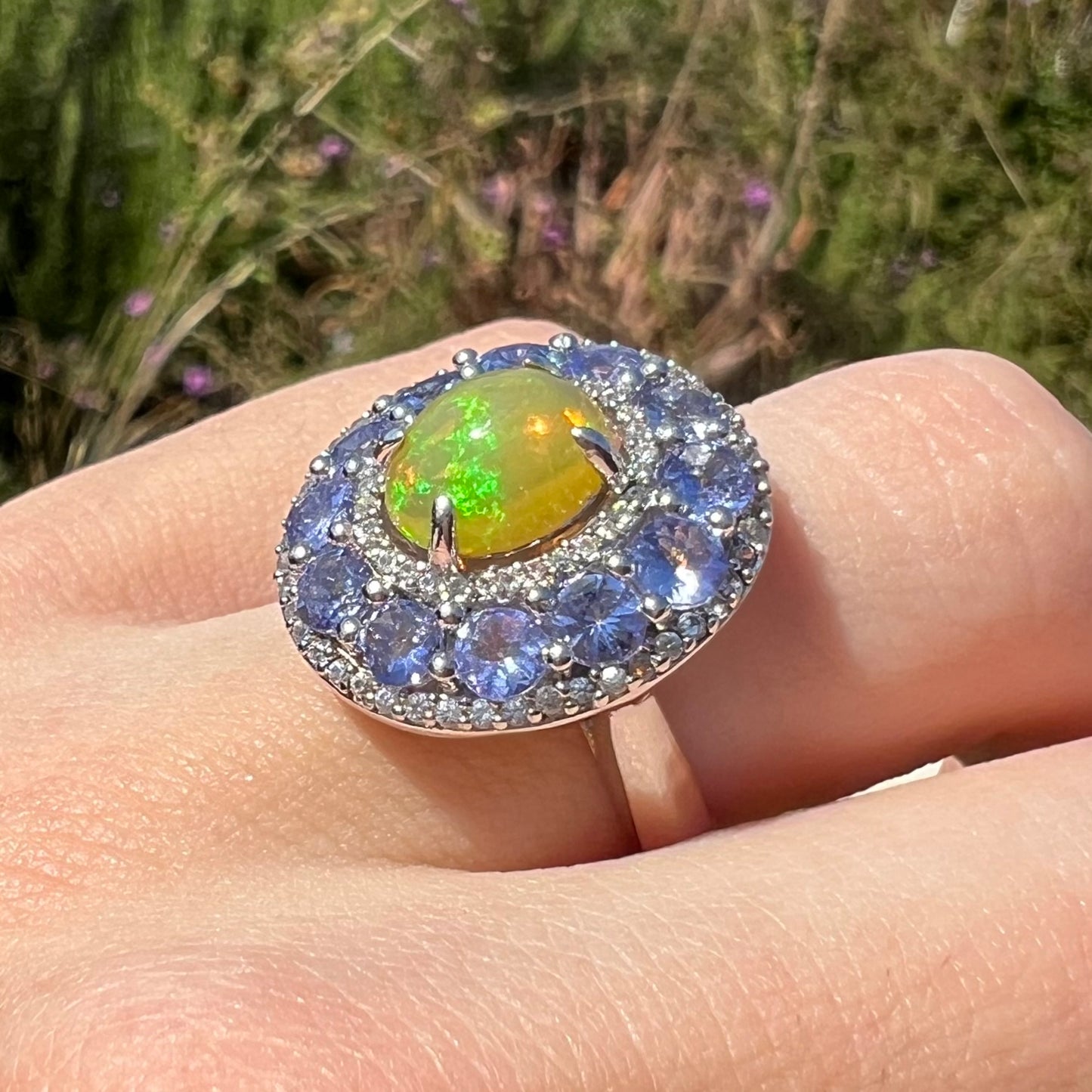 Ethiopian opal and tanzanite halo ring worn on a hand in the sunlight, shown at an angle. Opal shows green colors.