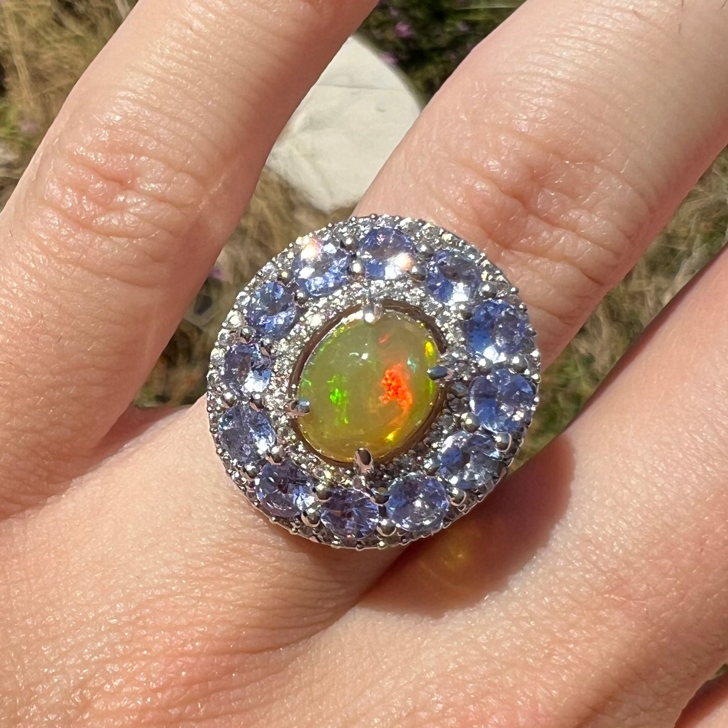 Macro front view of Ethiopian opal ring with tanzanite halo worn on hand in sunlight. Red colors seen in opal.