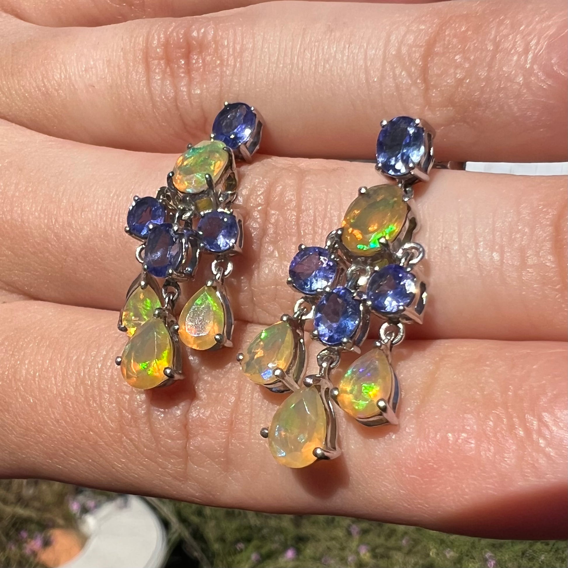 A pair of dangling sterling silver Ethiopian opal and tanzanite chandelier earrings, shown in sunlight at an angle.