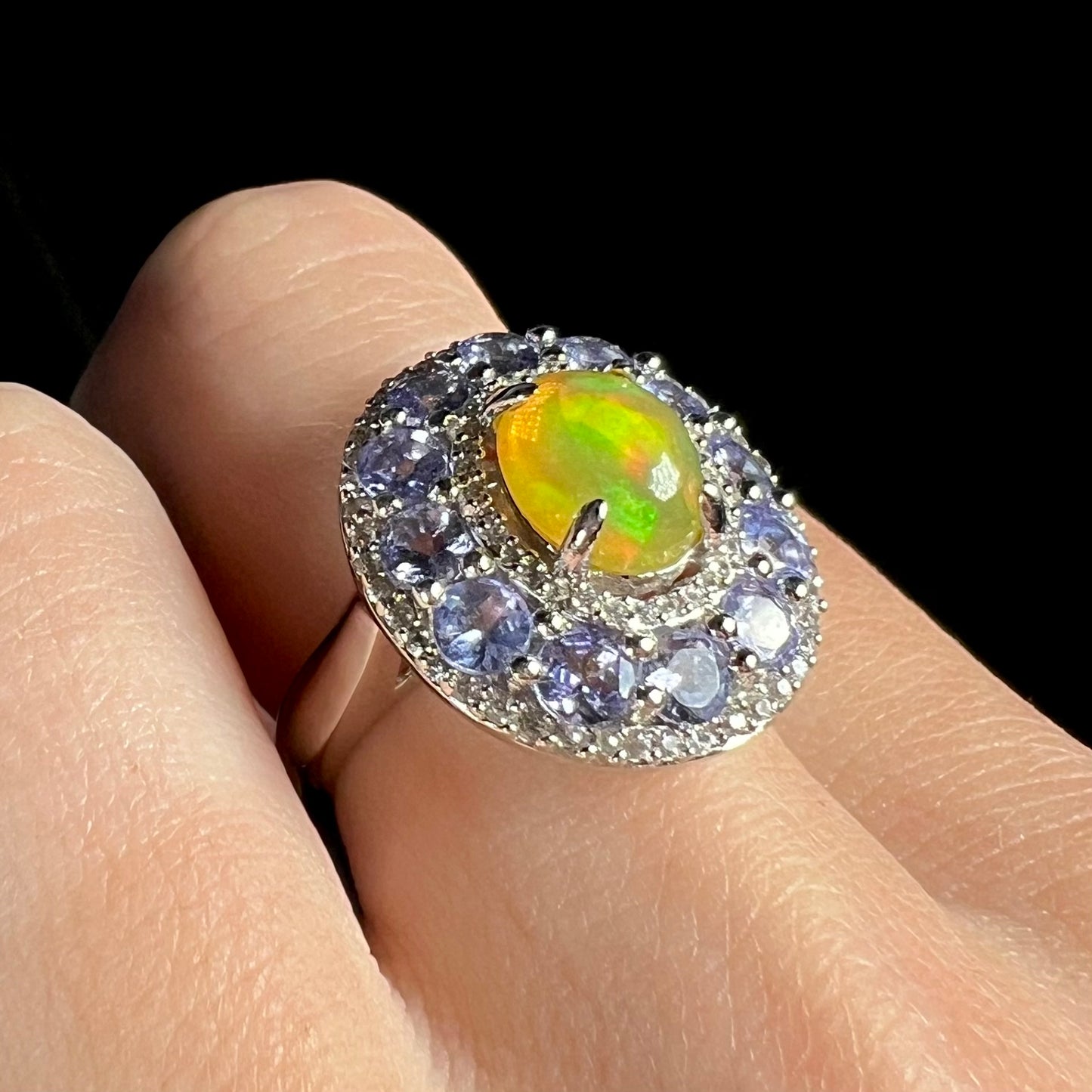 Ethiopian opal ring with tanzanite and white zircon halos worn on hand against black background.