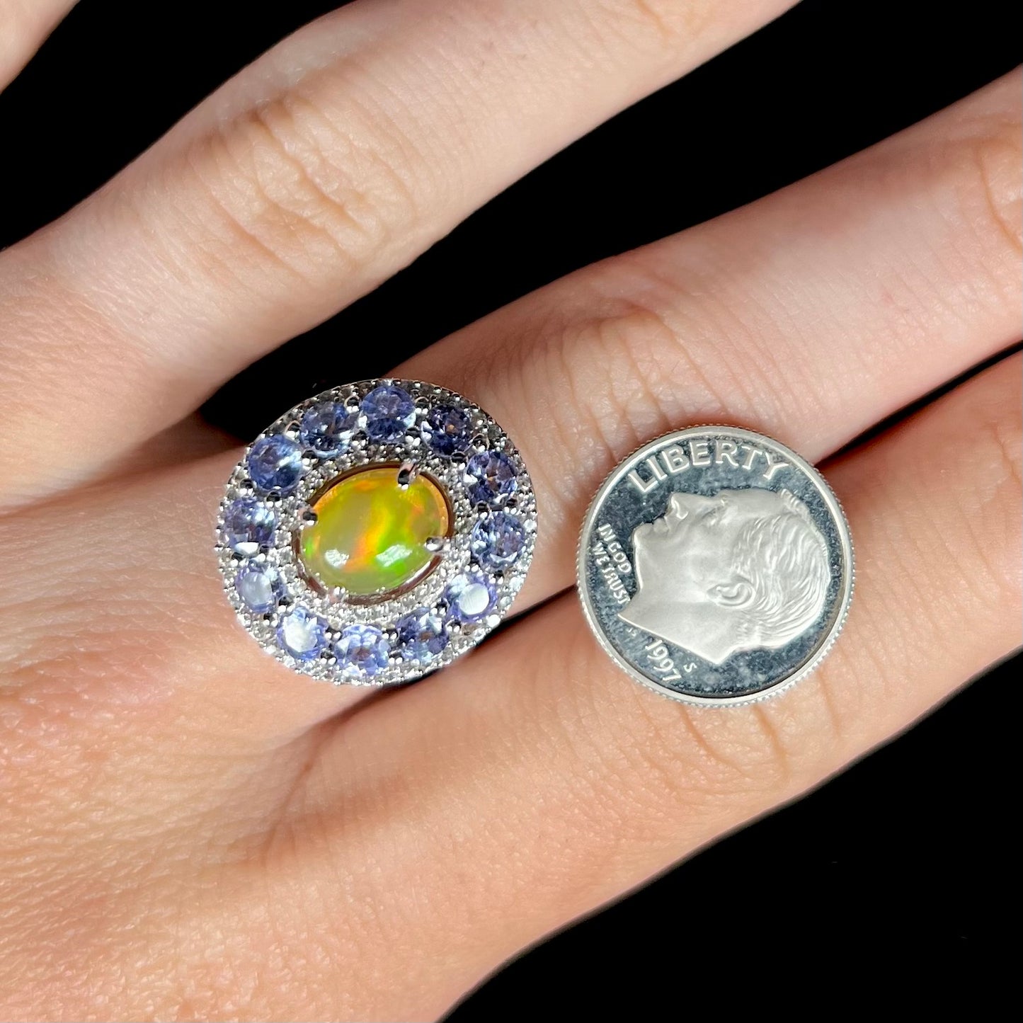 Ethiopian opal ring with triple gemstone halo worn on hand next to dime coin for scale.