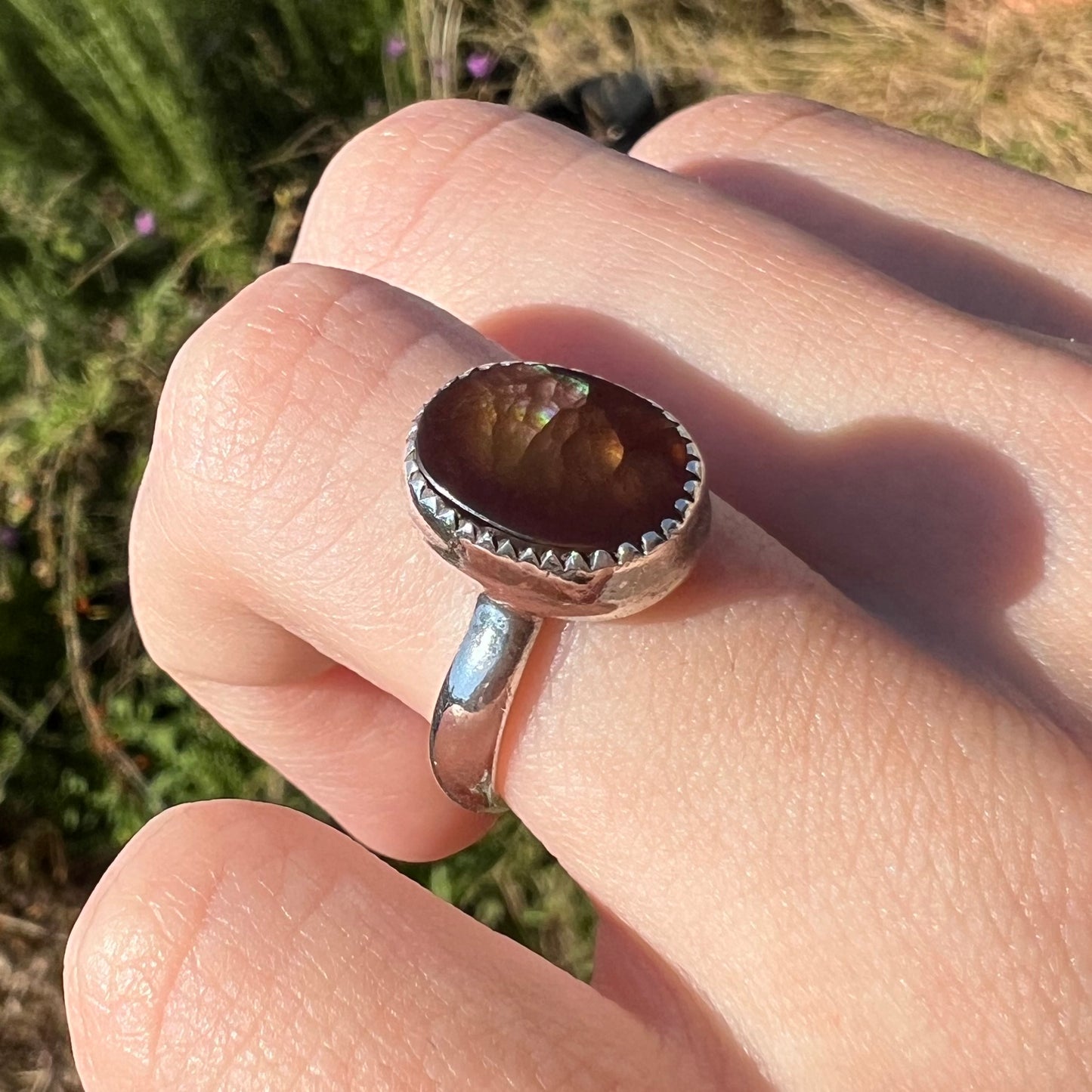 Silver solitaire ring with a brown fire agate stone on a person's finger against a natural background in sunlight.