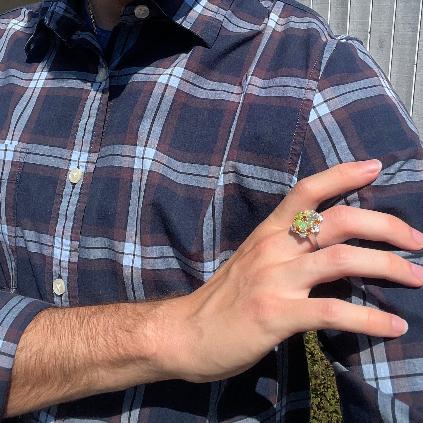 A person's hand resting on their shoulder wearing a fire opal cluster ring in the sunlight.