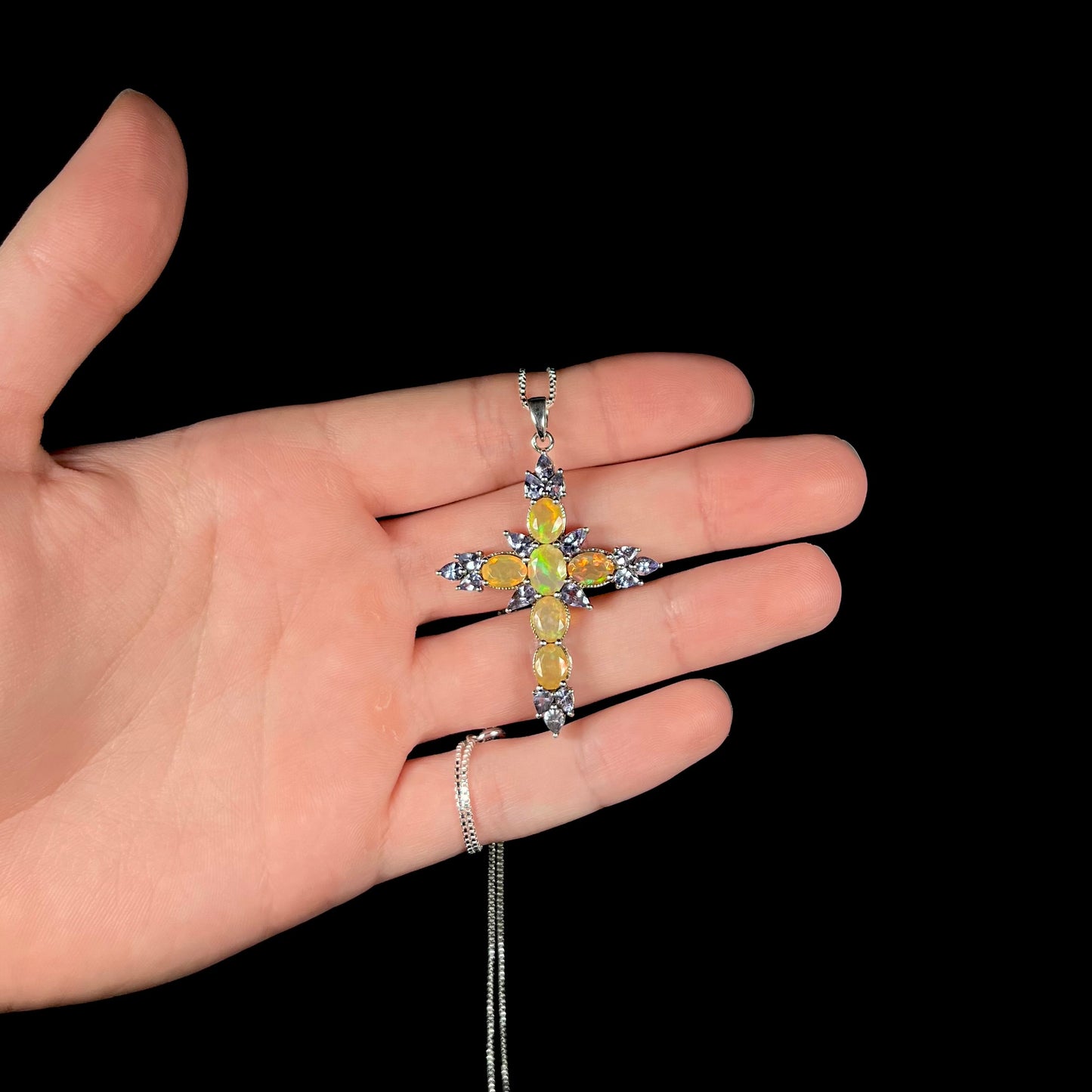 A fire opal cross necklace with tanzanite embellishments, held in a hand against a black background.