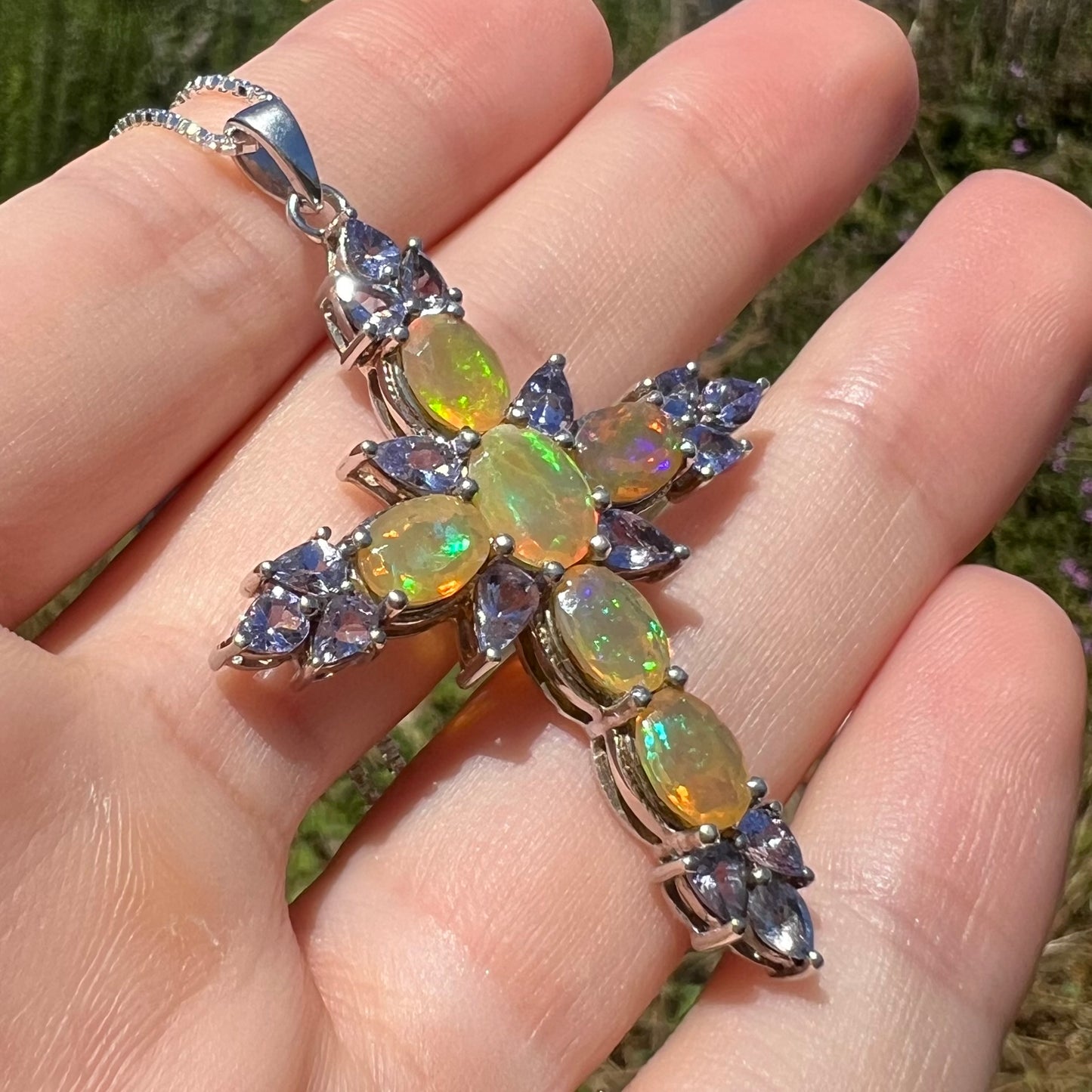 Angled close-up vof a fire opal cross pendant with tanzanite accents, held in sunlight.