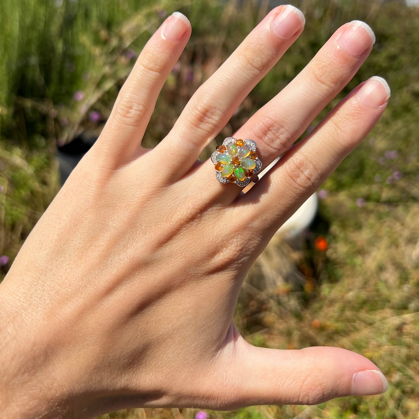 A hand wearing a fire opal and diamond flower-design ring in the sunlight against blurred outdoor background.