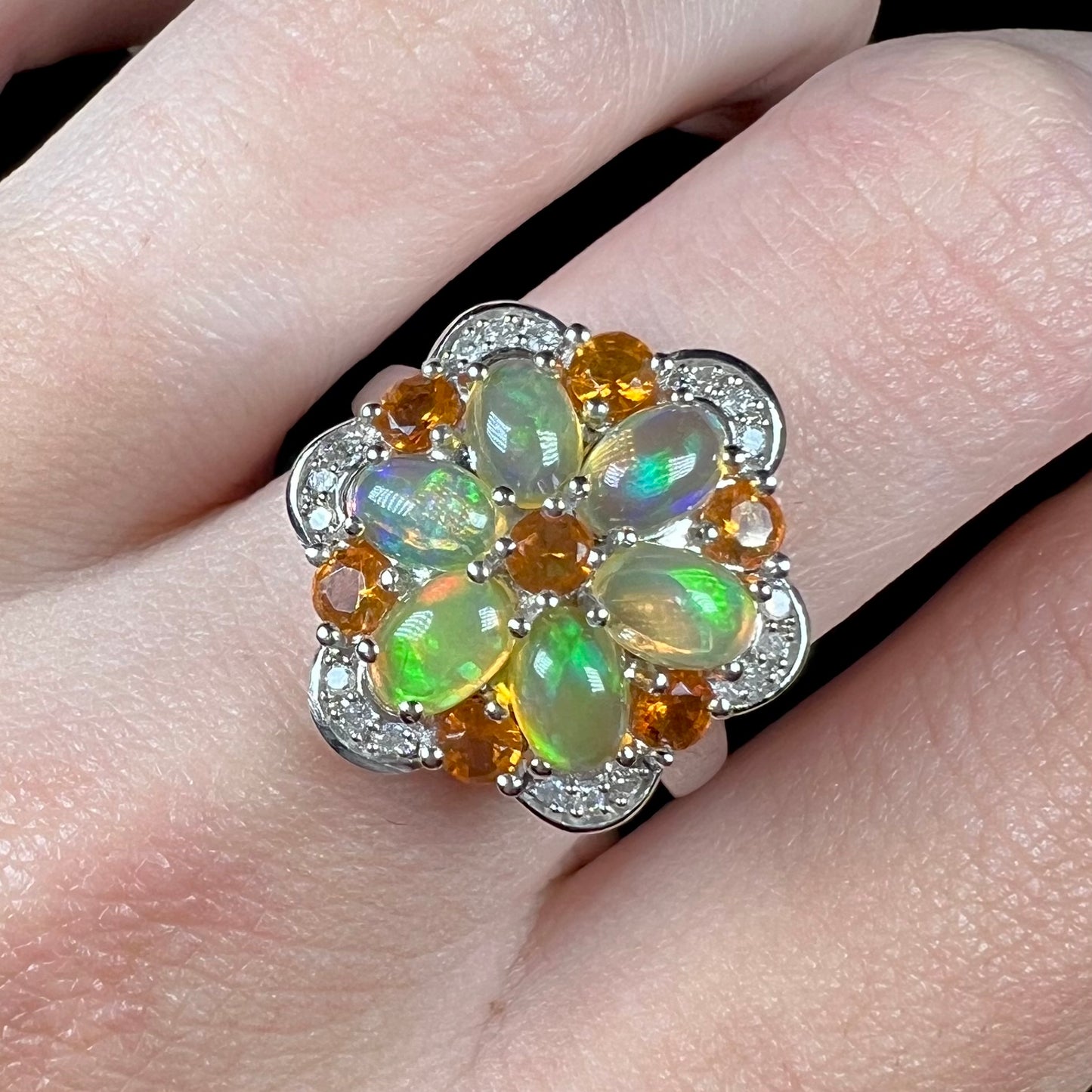 Front macro view of fire opal and diamond flower ring worn on a finger against a black back backdrop.