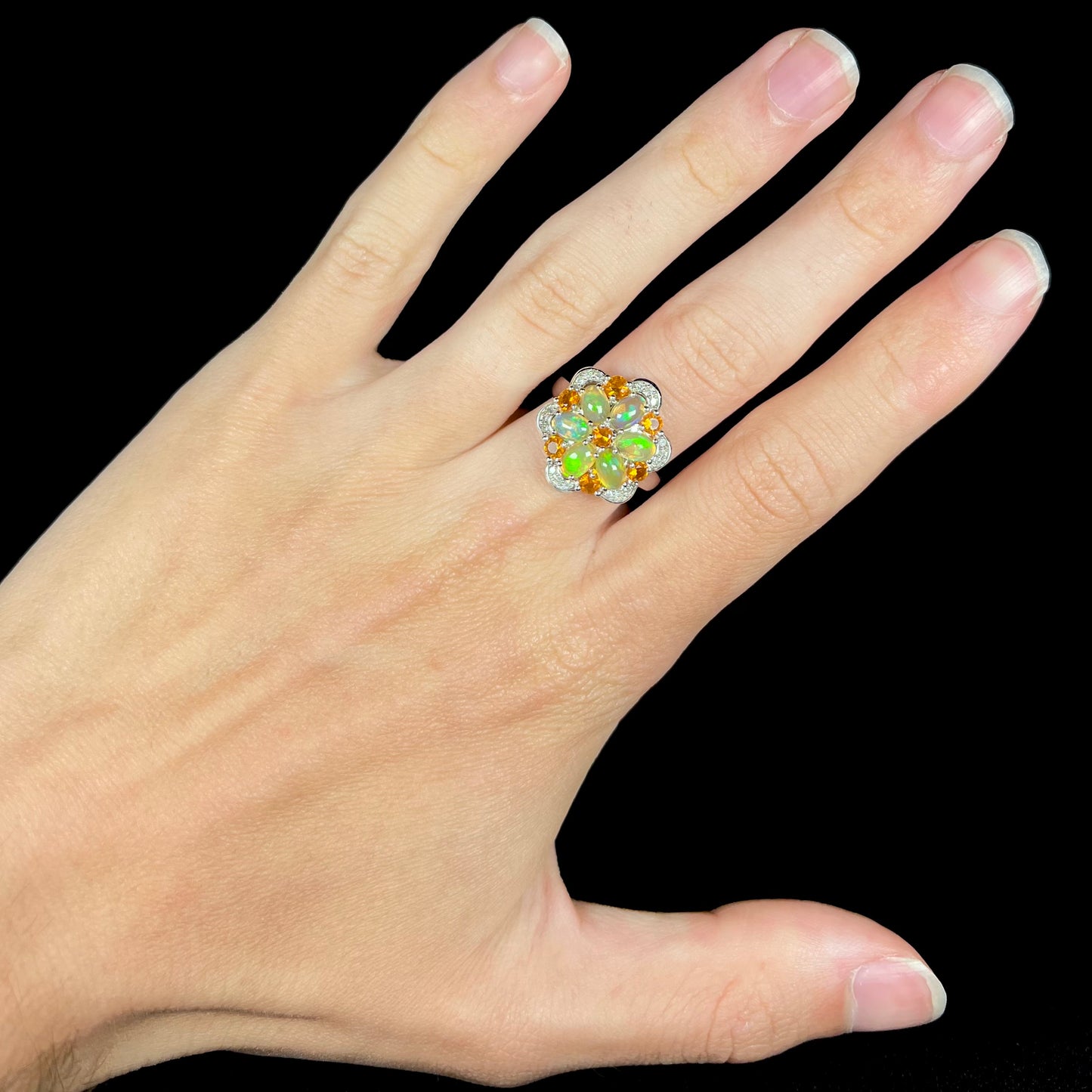 A fire opal flower ring with diamonds, worn on a hand against a black backdrop.