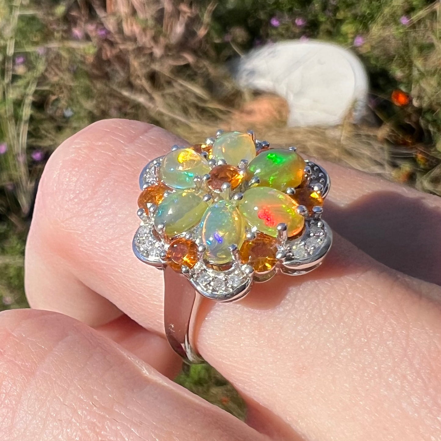 A fire opal flower ring worn on a finger in the sunlight at an angle against a blurred natural backdrop.