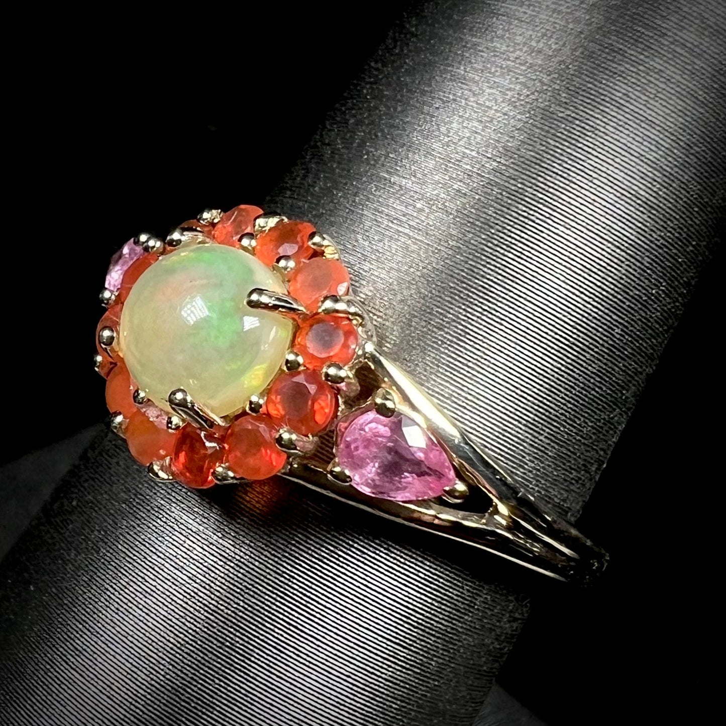 A yellow gold ring mounted with a round Ethiopian opal in a halo of faceted Mexican fire opals. Two pear shaped pink tourmalines accent the sides of the ring.