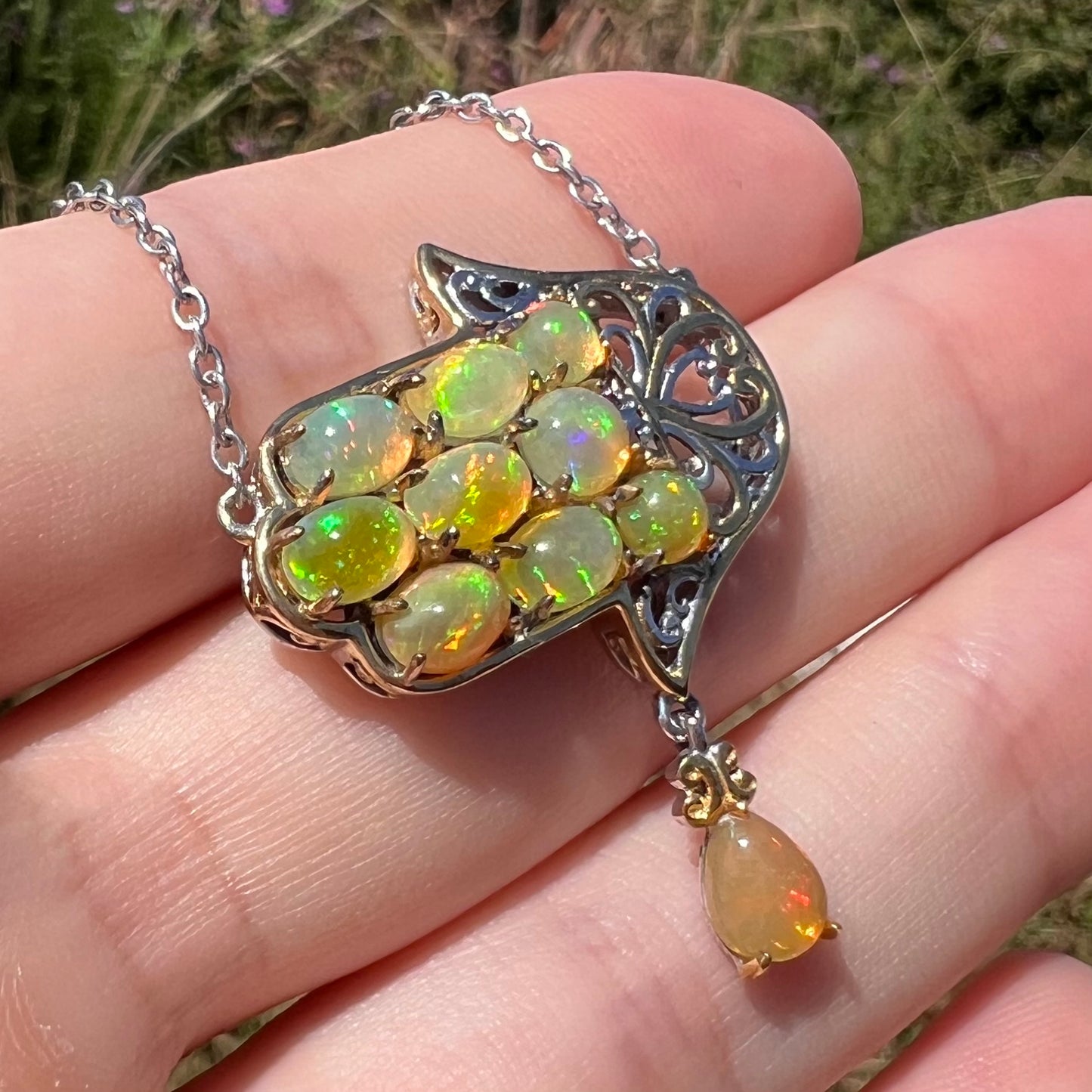 A fire opal hamsa necklace set with Ethipoian opals, shown in a hand in the sunlight.