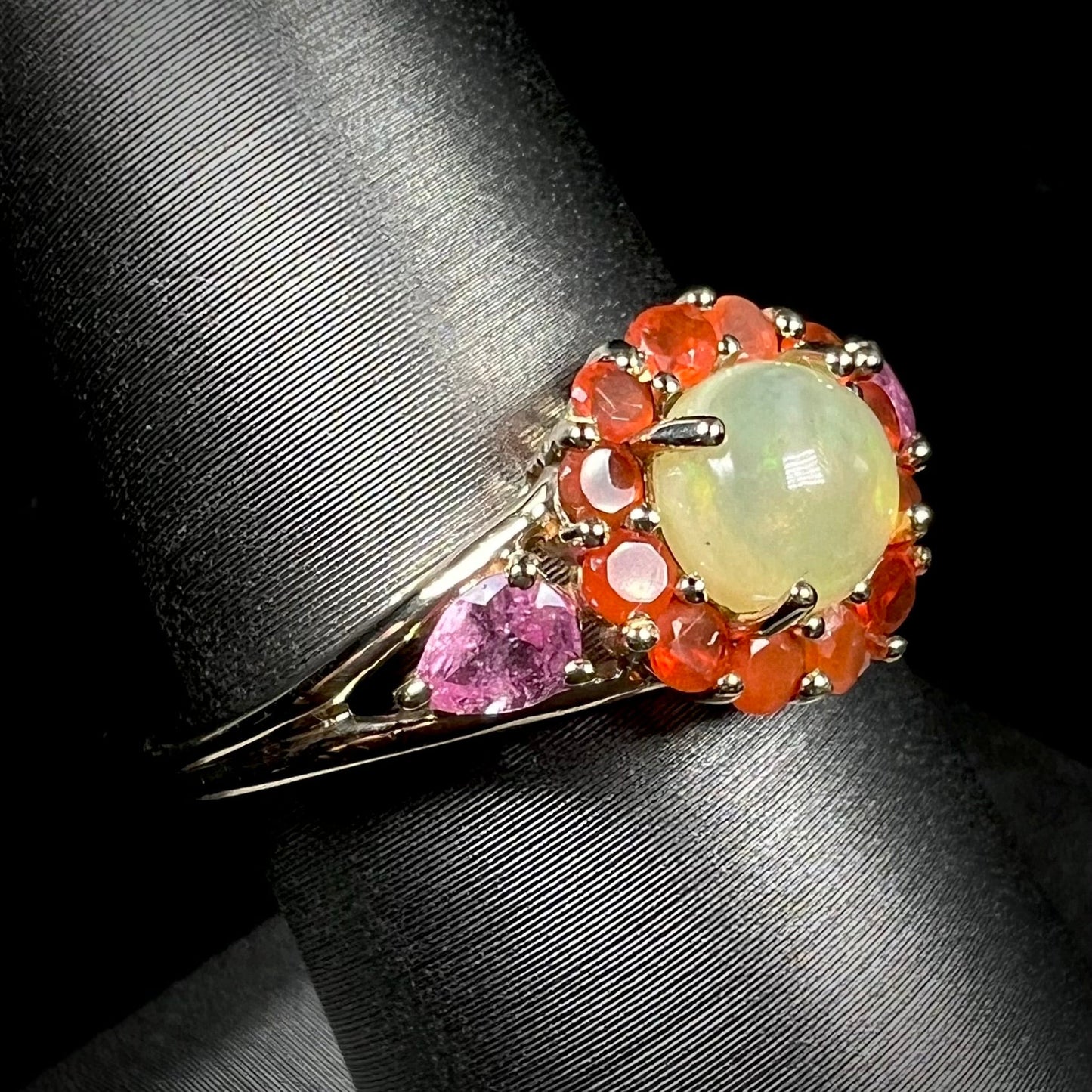 A yellow gold ring mounted with a round Ethiopian opal in a halo of faceted Mexican fire opals. Two pear shaped pink tourmalines accent the sides of the ring.