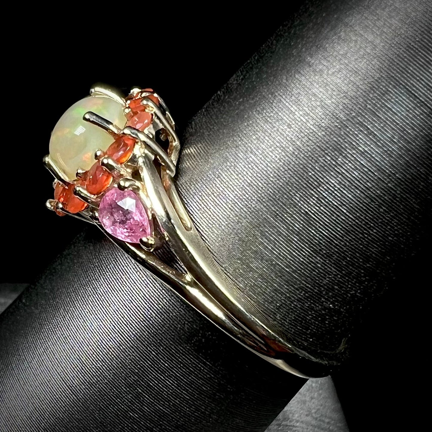 A yellow gold ring mounted with a round Ethiopian opal in a halo of faceted Mexican fire opals. Two pear shaped pink tourmalines accent the sides of the ring.
