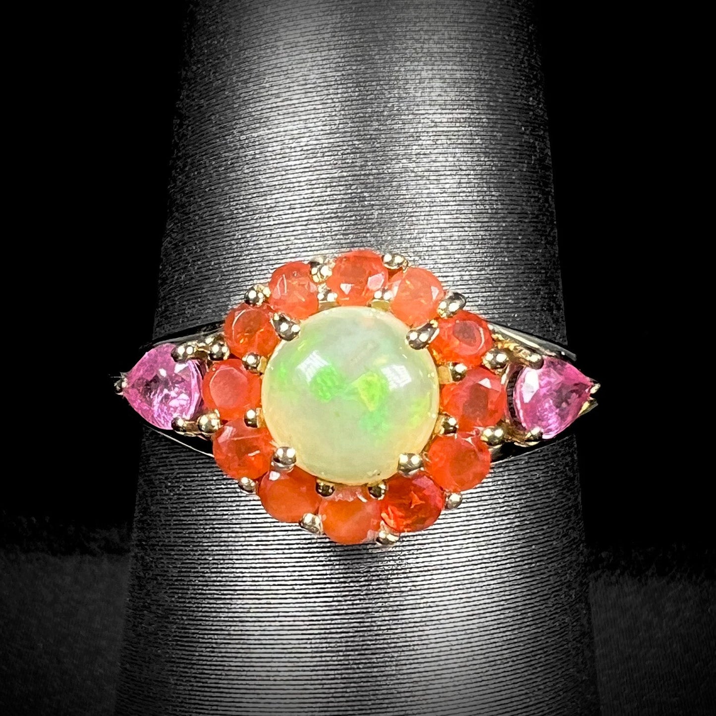 A yellow gold ring mounted with a round Ethiopian opal in a halo of faceted Mexican fire opals. Two pear shaped pink tourmalines accent the sides of the ring.