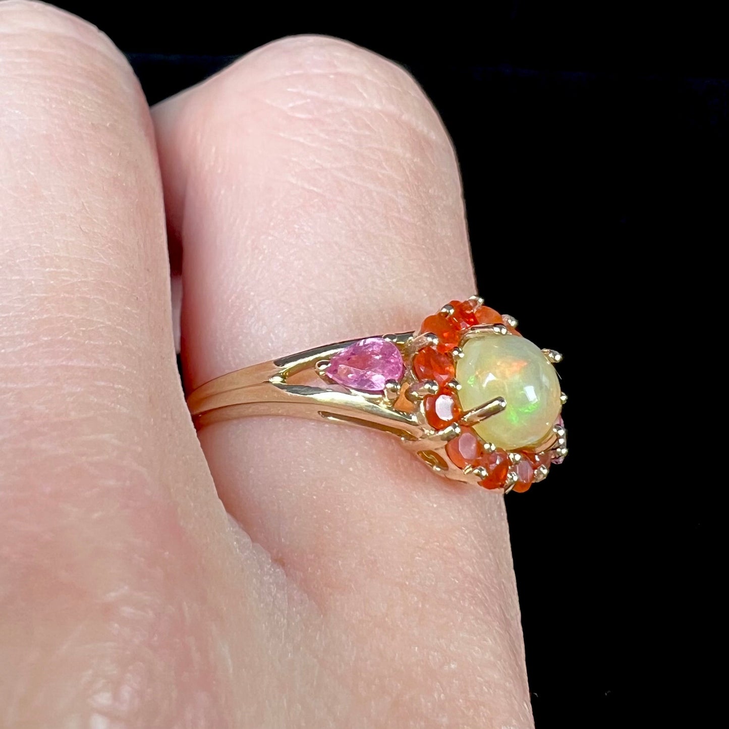 A yellow gold ring mounted with a round Ethiopian opal in a halo of faceted Mexican fire opals. Two pear shaped pink tourmalines accent the sides of the ring.