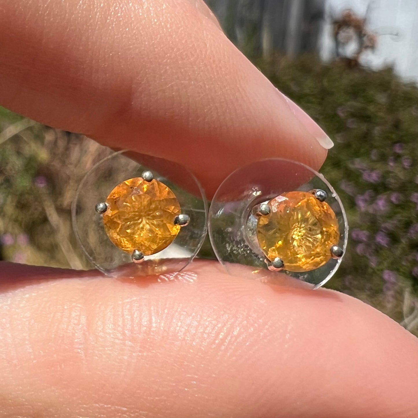 Close-up front view of Mexican fire opal stud earrings in sterling silver, held between two fingers in sunlight.