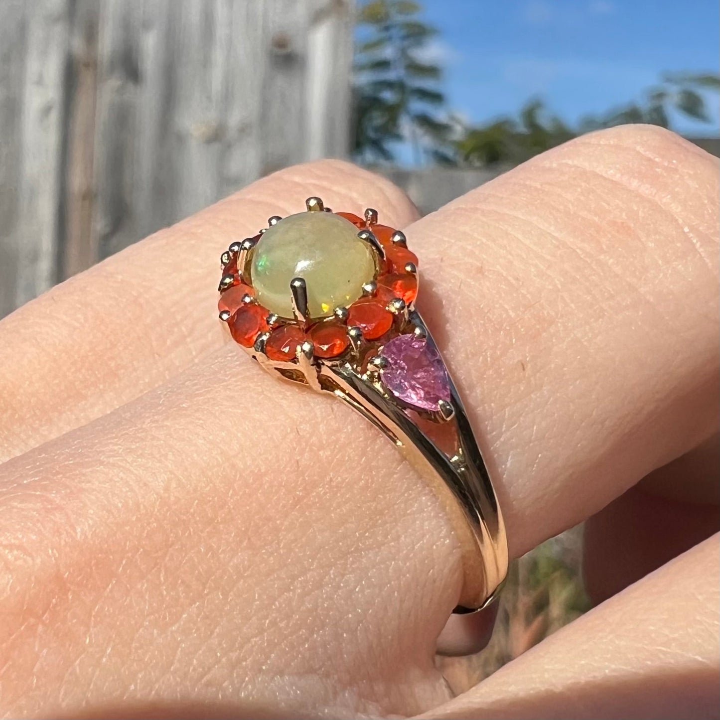 A yellow gold ring mounted with a round Ethiopian opal in a halo of faceted Mexican fire opals. Two pear shaped pink tourmalines accent the sides of the ring.