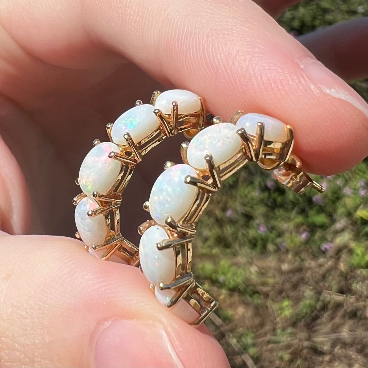 Pair of five-stone natural Australian opal semi-hoop earrings in 14k yellow gold shown from angled close-up, held between fingers in sunlight