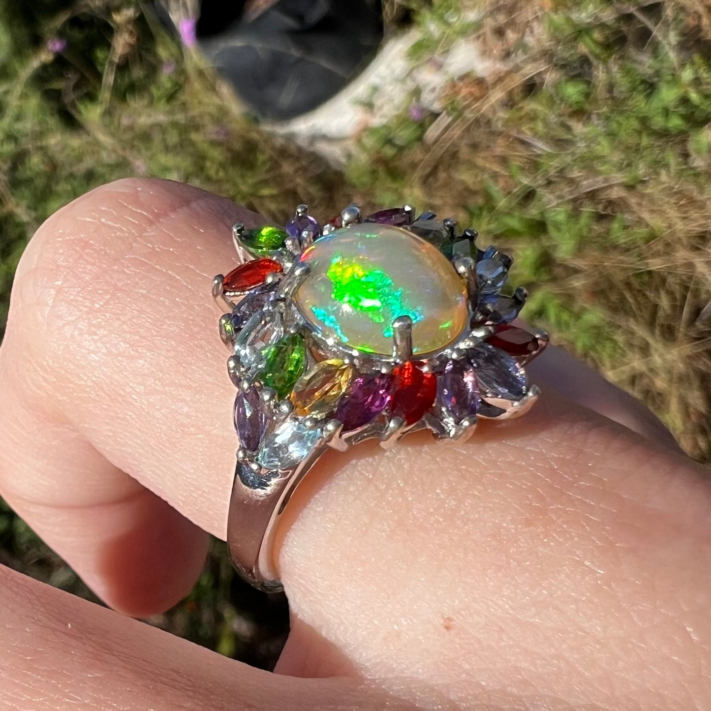 A flamboyant opal and gemstone ring shown in daylight, worn on a finger at an angle. A rainbow of colors are seen.