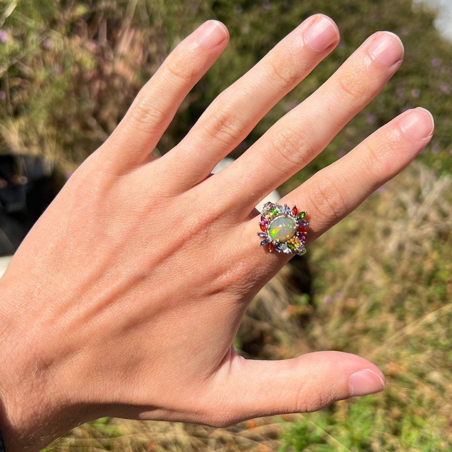 A flamboyant style opal ring with multicolor gemstone accents, worn on a hand at a distance in sunlight.