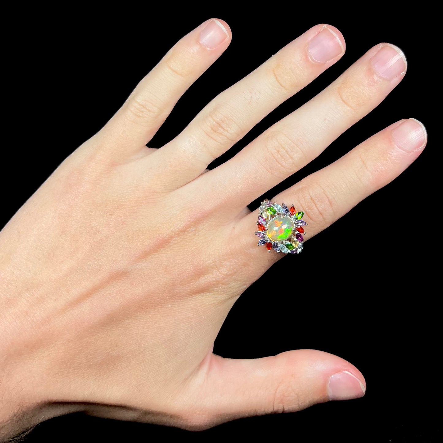 A flamboyant style opal and multi-gemstone ring, worn on a hand against a black background.