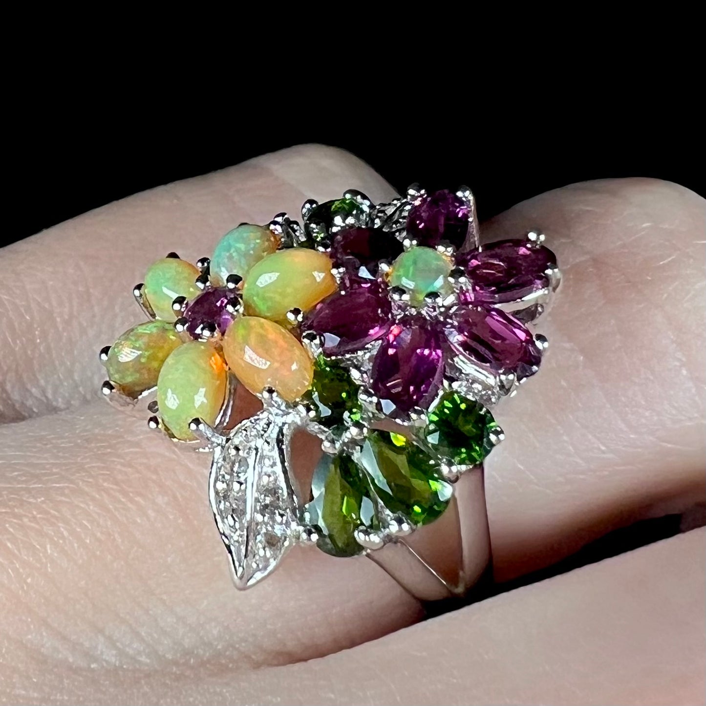 A flower design opal ring with garnet and chrome diopside accents, worn on a finger and shown at an angle.