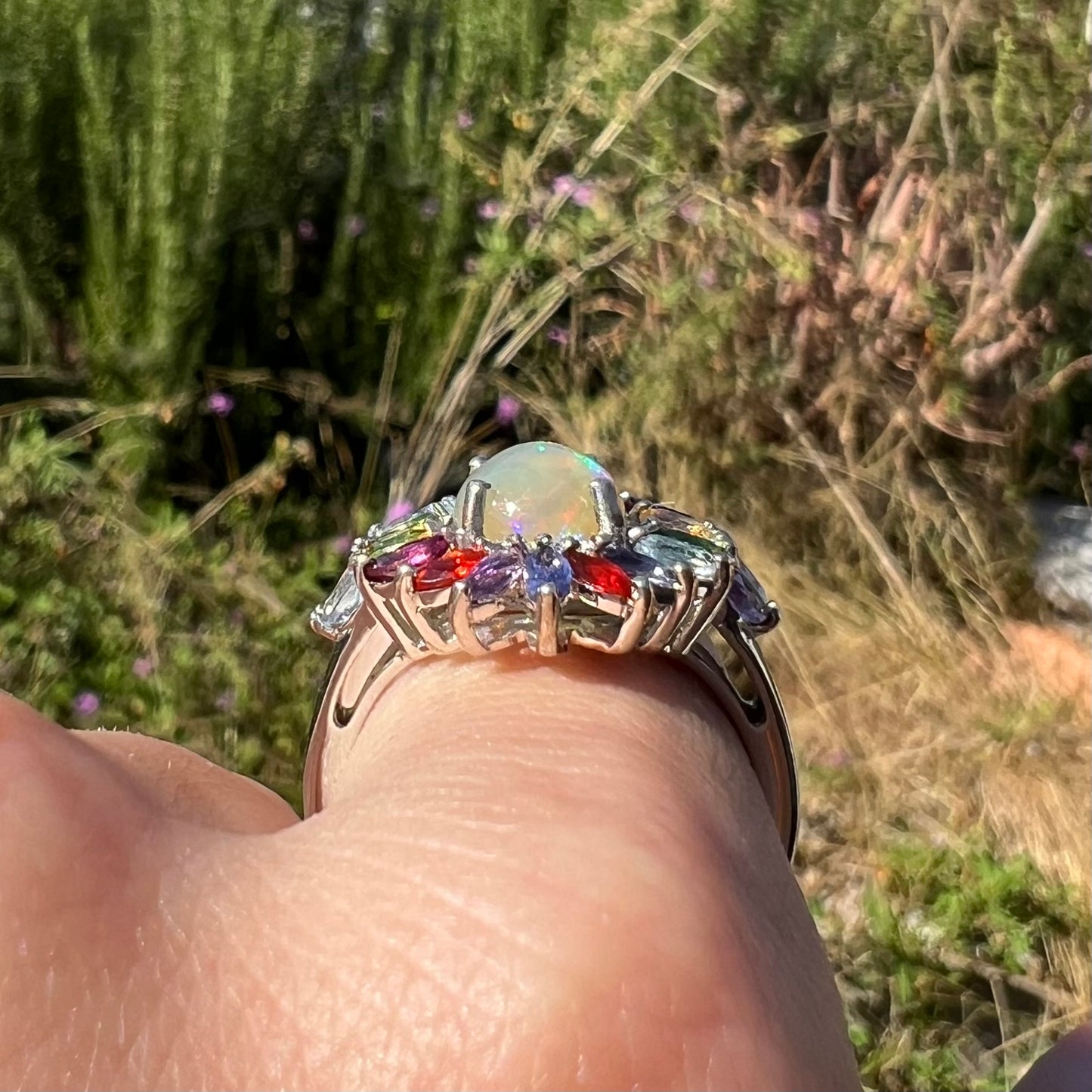 Side gallery view of an opal and gemstone ring made from silver on a hand in sunlight.