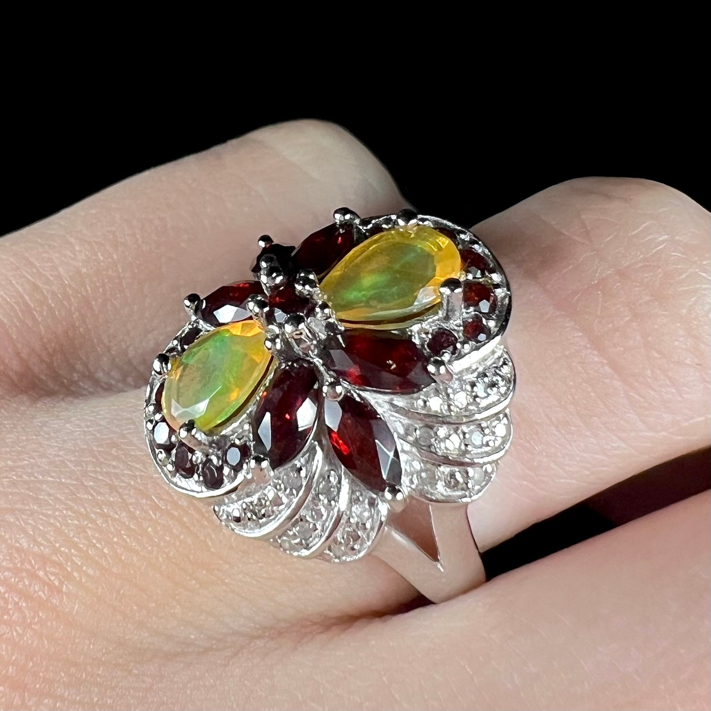 A sterling silver Ethiopian opal and garnet infinity-design ring with diamond accents, shown at an angle on a finger.