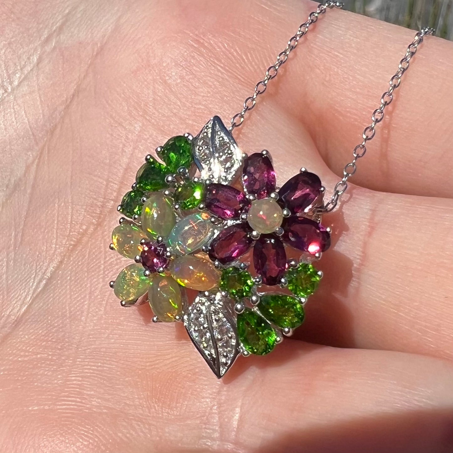 A sterling silver opal and garnet flower necklace shown close-up in the palm of a hand at an angle in sunlight.