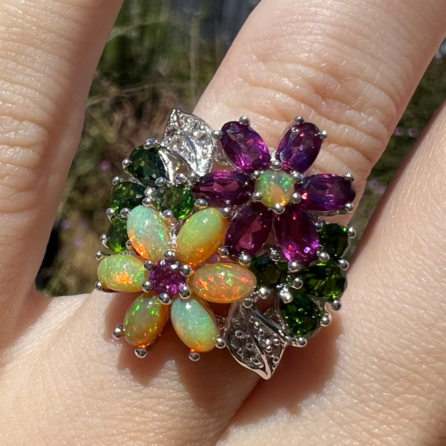 Close-up front view of a gemstone flower ring with Ethiopian opal and garnet, worn on a finger in sunlight.