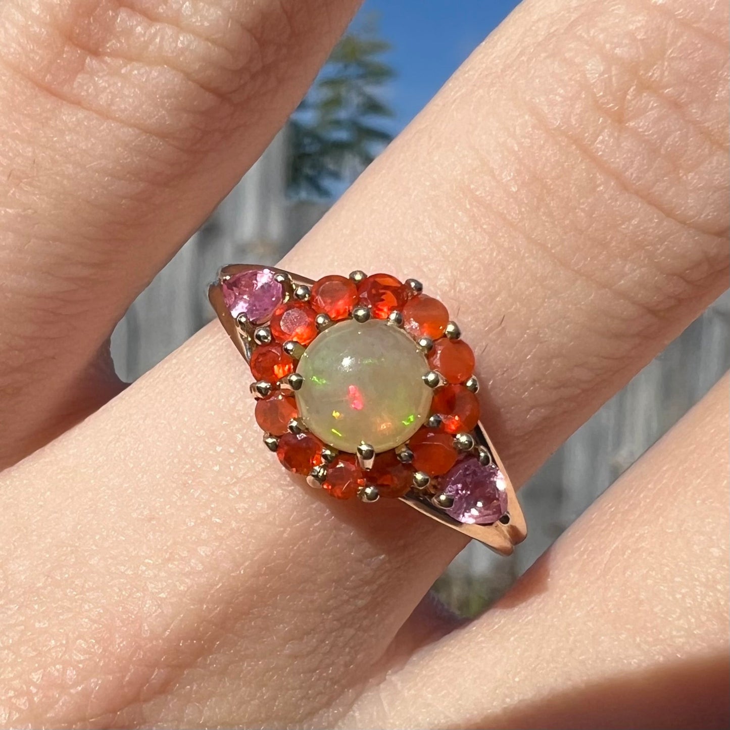 A yellow gold ring mounted with a round Ethiopian opal in a halo of faceted Mexican fire opals. Two pear shaped pink tourmalines accent the sides of the ring.