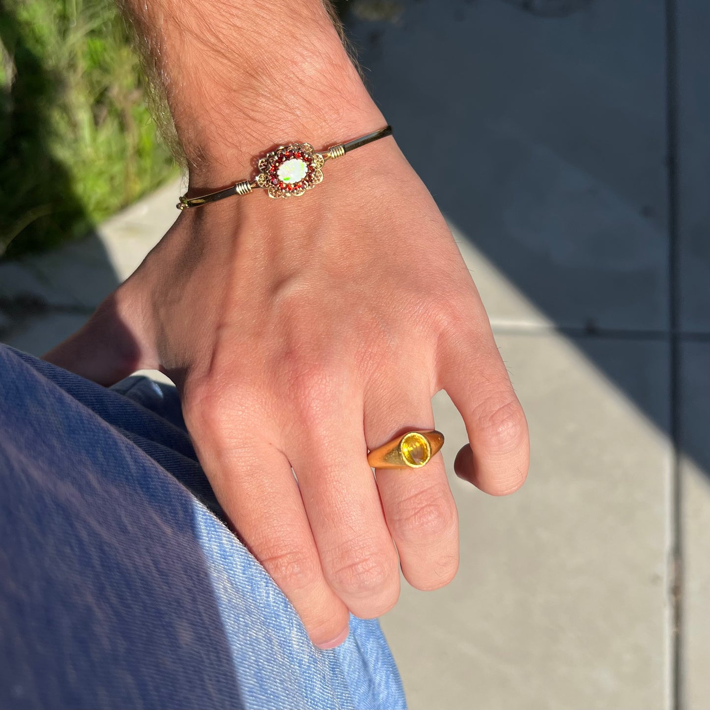 A vintage yellow gold opal and garnet halo bangle bracelet shown on a wrist in sunlight, hand resting on hip.