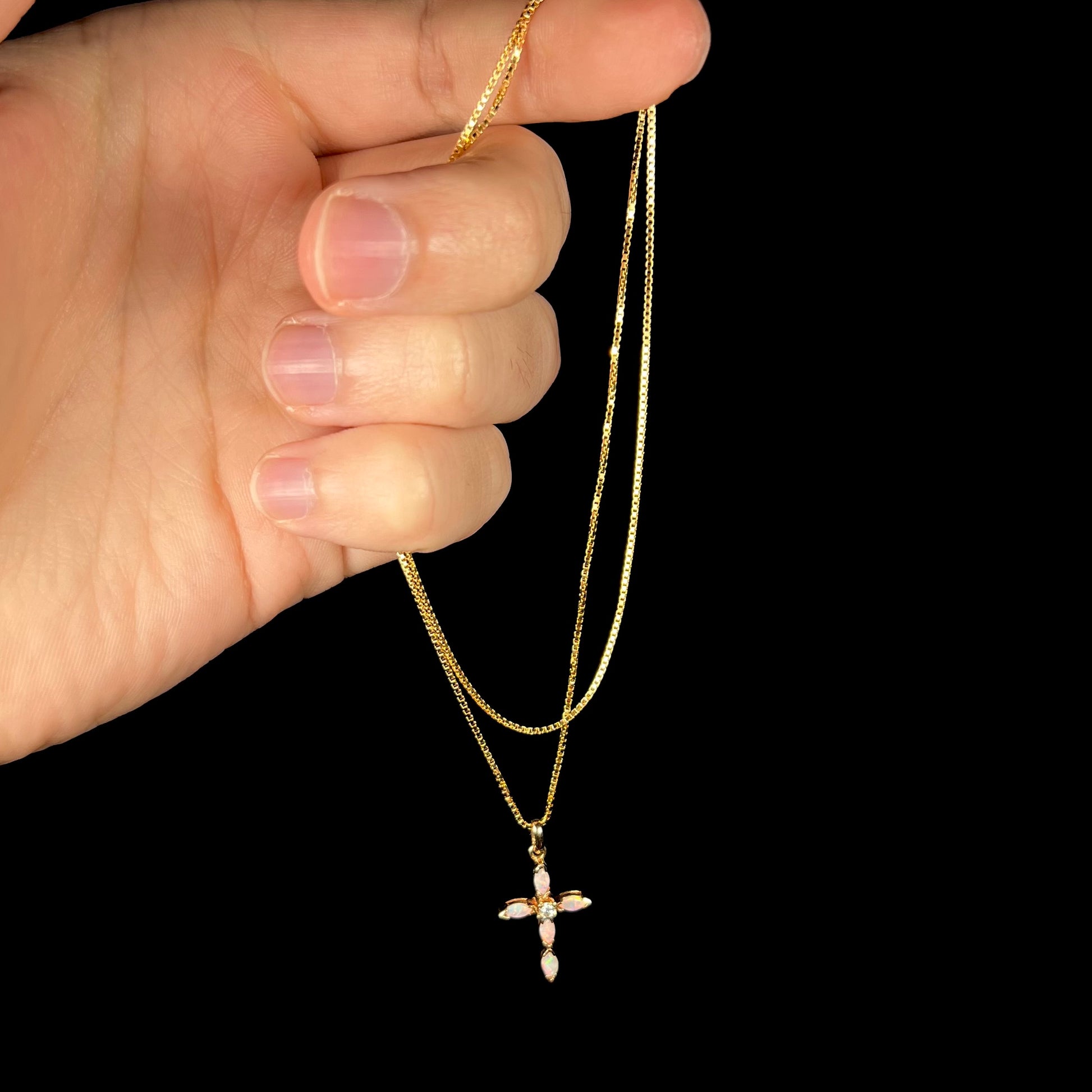 A gold opal cross necklace dangled from a hand by the chain against a black background.