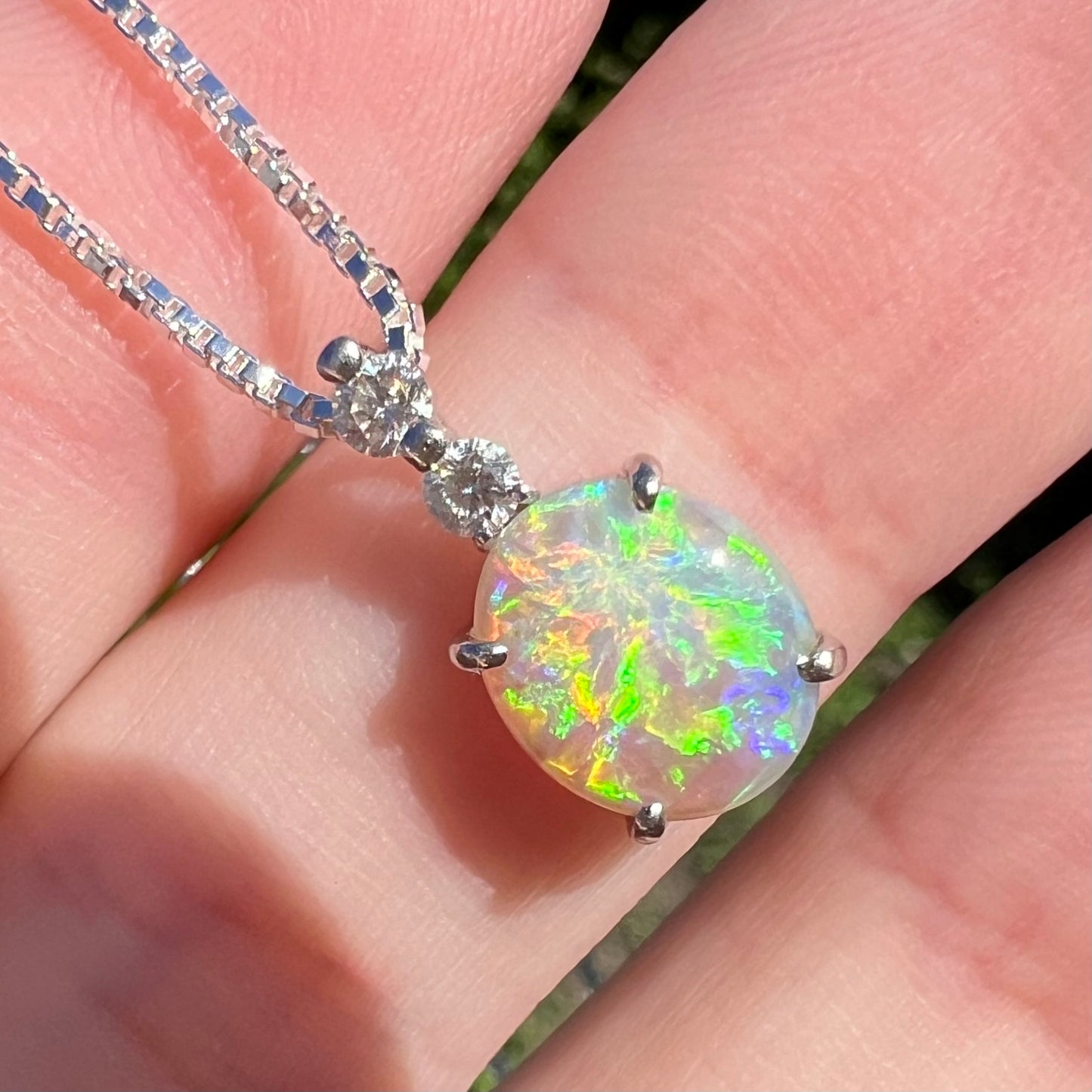 Close-up macro shot of a dark opal pendant in sunlight against a finger, displaying vivid green and orange flashes.