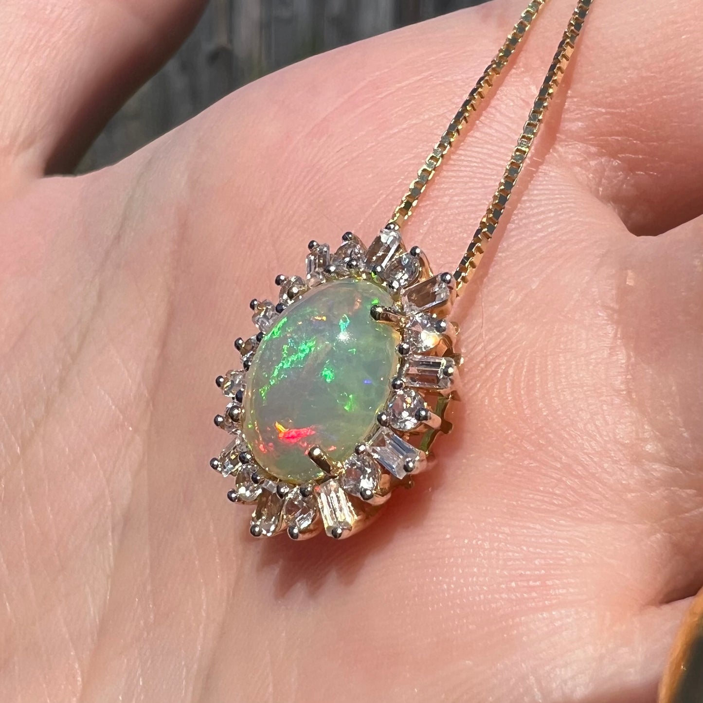 Green and red fire are seen in yellow gold Ethiopian opal pendant featuring white sapphire halo design. Pendant is seen from angle in sunlight, held in hand.