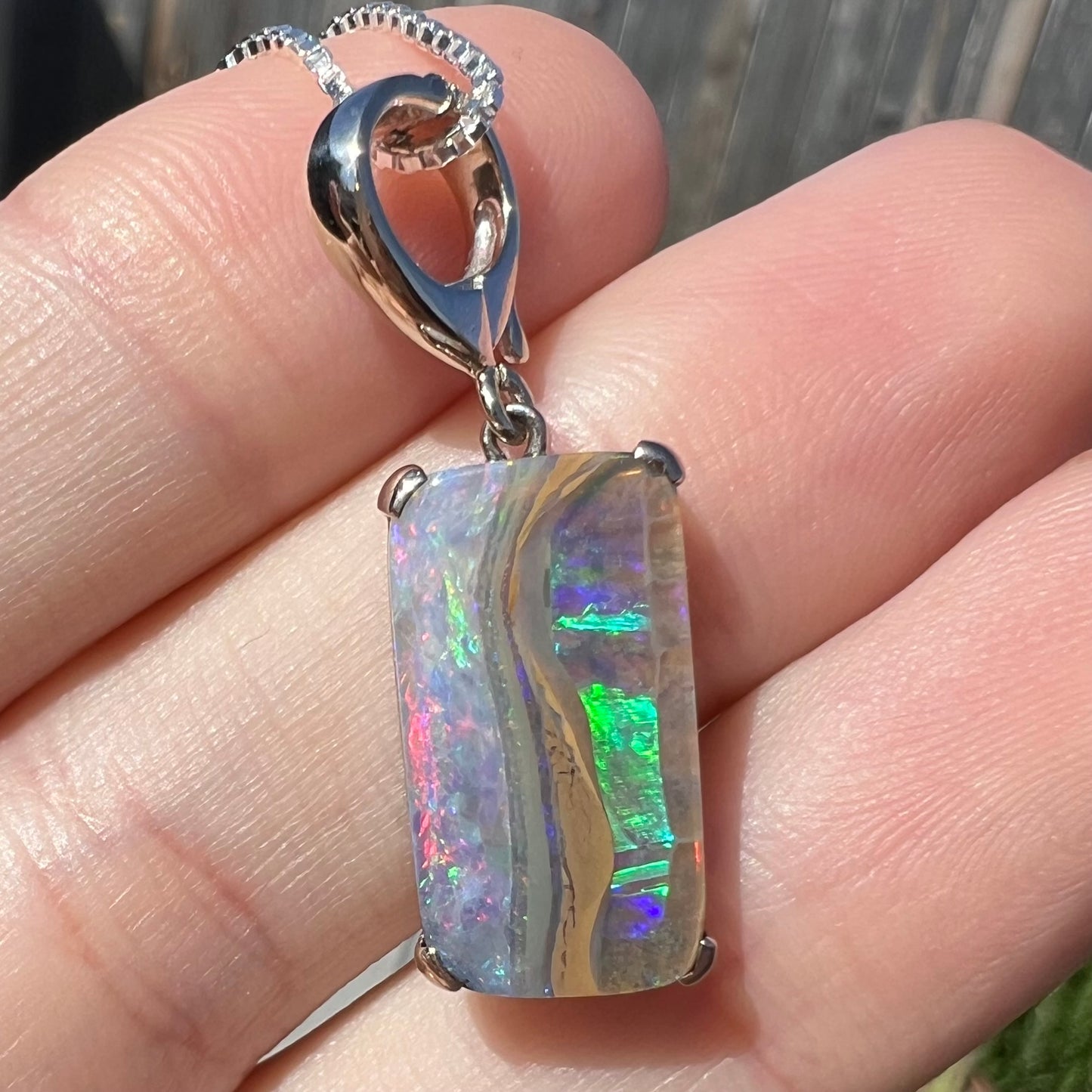 Macro close-up of the Hakumei boulder opal pendant against a finger in sunlight, with blues and greens dominating one side and pinks glowing on the other.