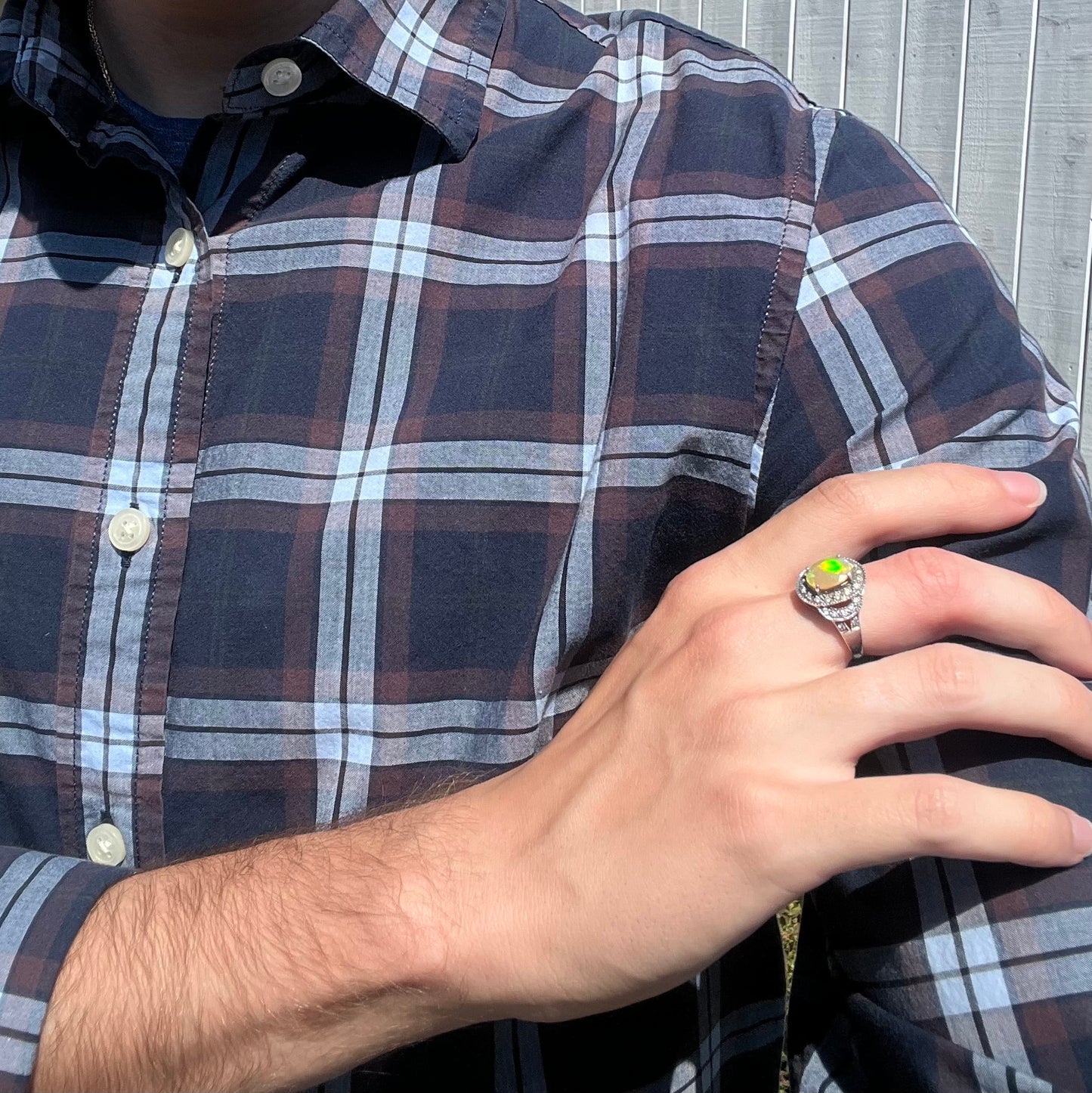 Person wearing a plaid shirt crossing their arm to hold their shoulder while wearing a halo-style opal ring.