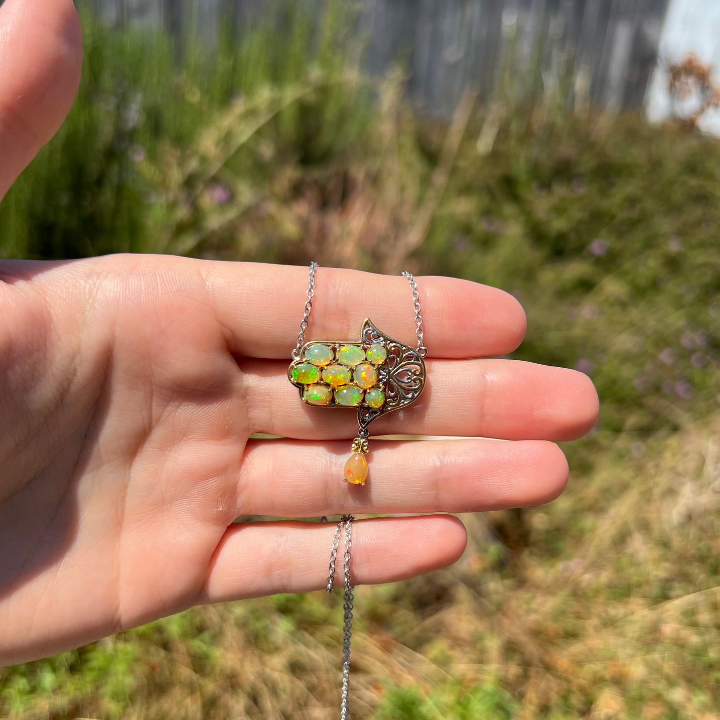 An hamsa opal necklace held in a hand at a distance in the sunlight.