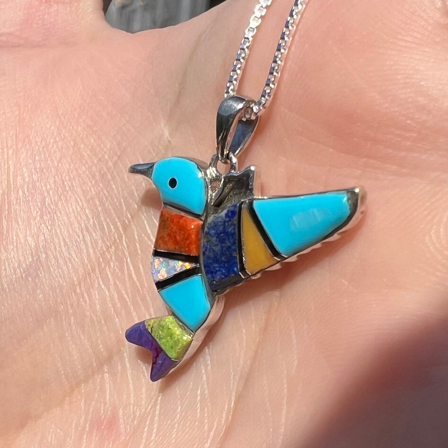 A multi-stone inlay hummingbird necklace in sterling silver, shown at an angle close-up in a hand under sunlight.