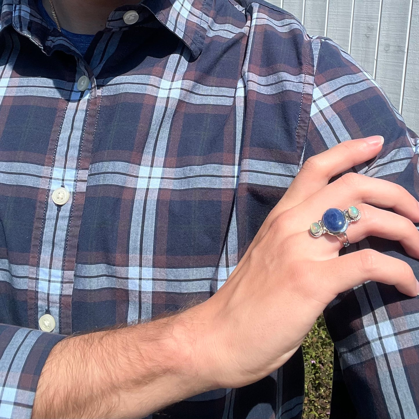 A person with a blue plaid shirt holding their hand on their shoulder, wearing a kyanite opal ring.