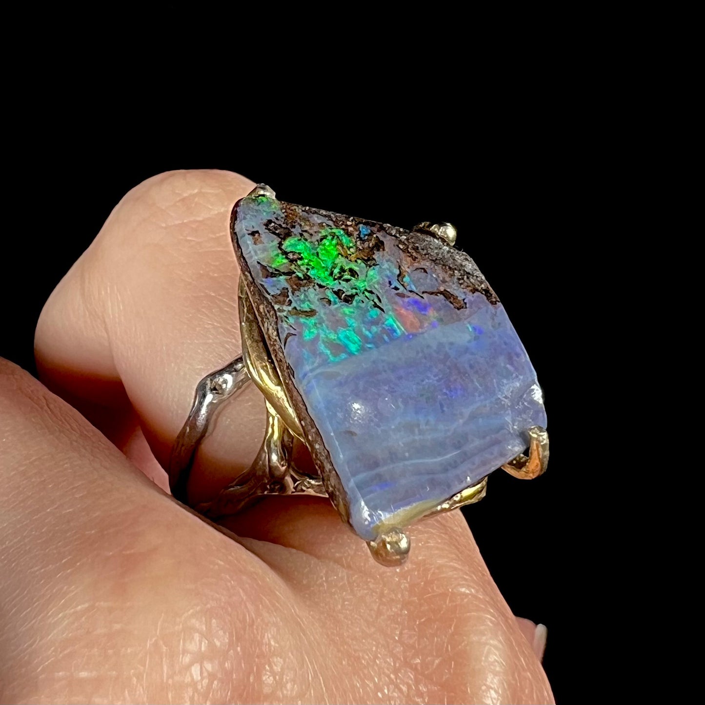 A large boulder opal ring worn on a finger, showing green and blue color flashes amid its ironstone matrix.