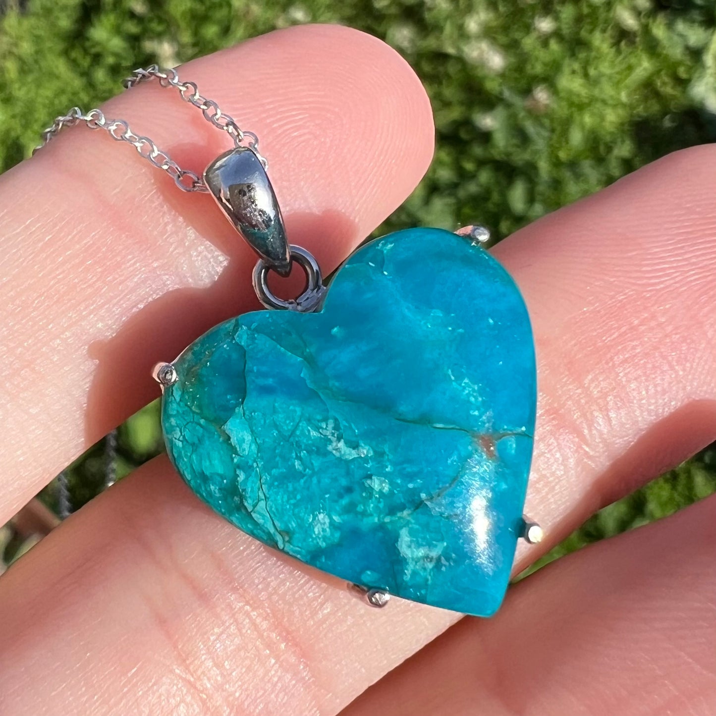 Macro straight-on view of vibrant blue Chilean opal heart necklace in sunlight, resting against fingers.