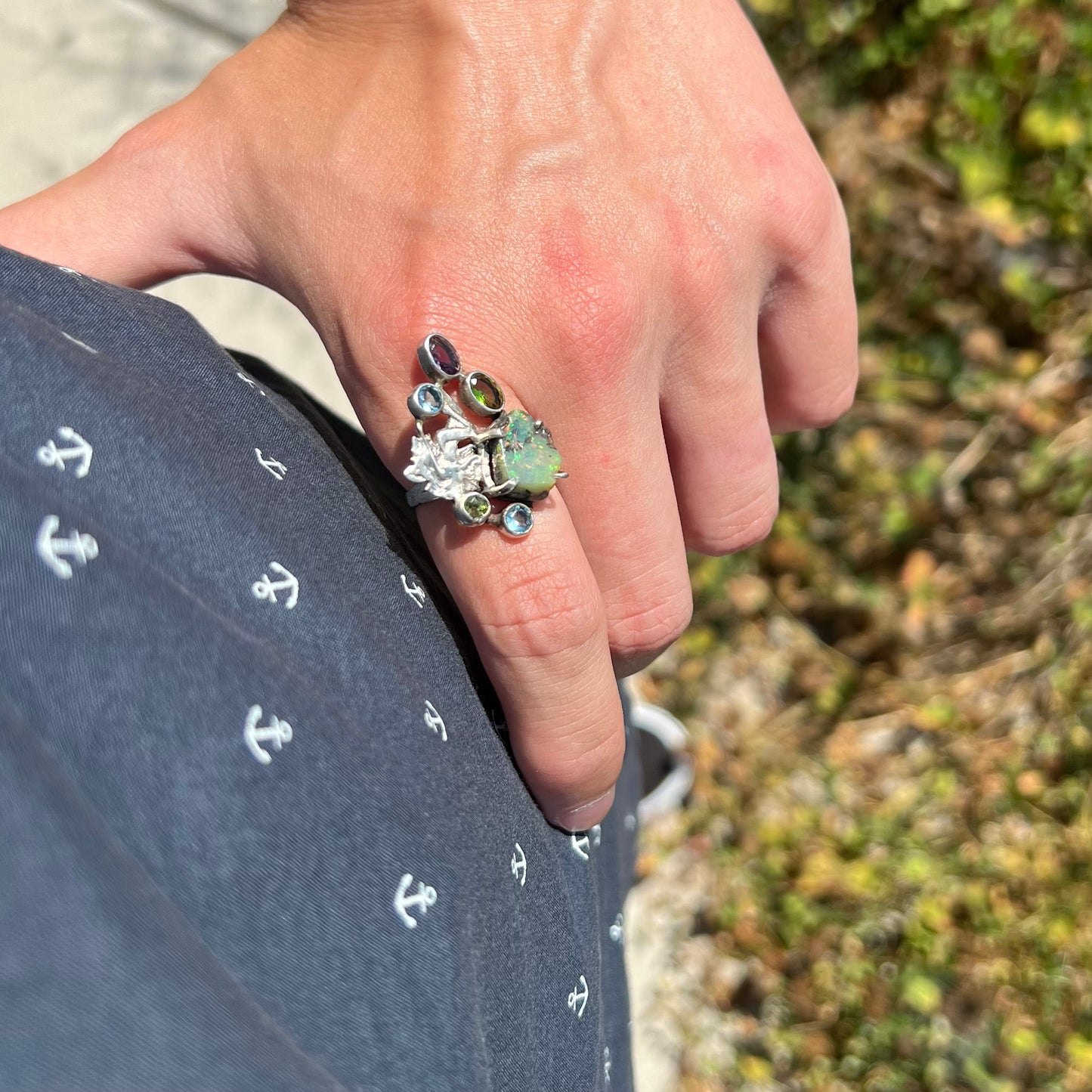 A silver men's ring set with rough Virgin Valley opal and gems with a female figure design, worn on a hand resting on a hip.