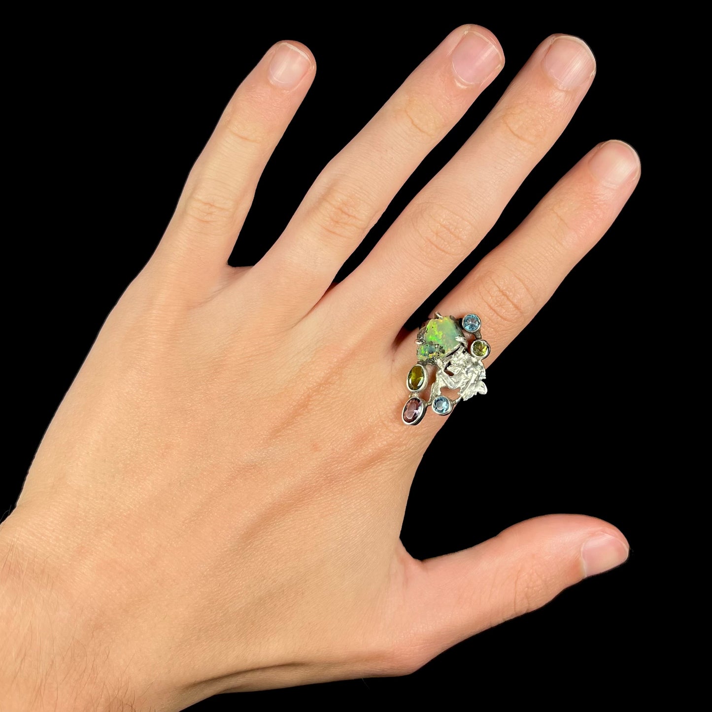 A silver men's ring set with rough Virgin Valley opal and gems with a female figure design worn on a hand.