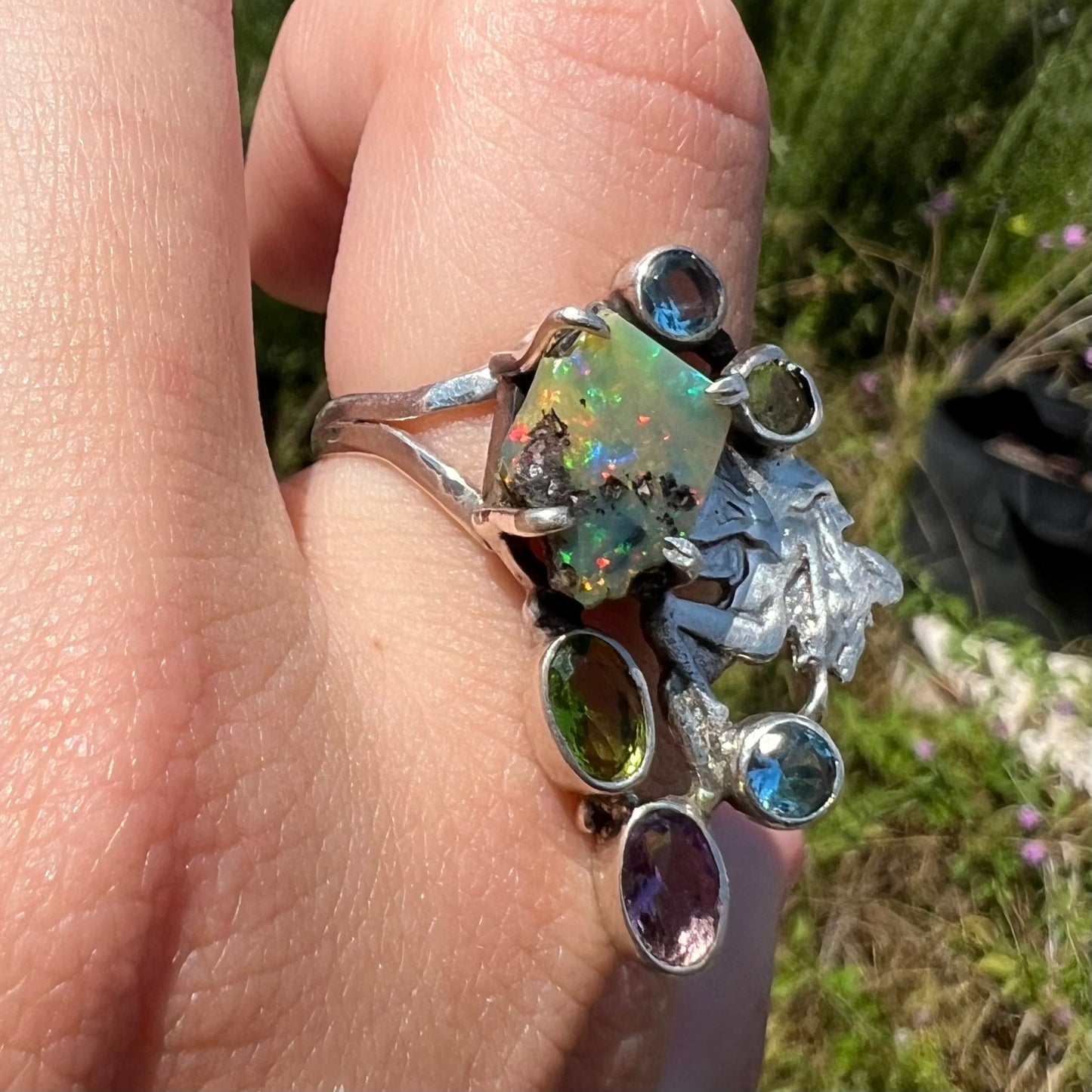 A silver men's ring set with rough Virgin Valley opal and gems with a female figure design shown in the sunlight at an angle.