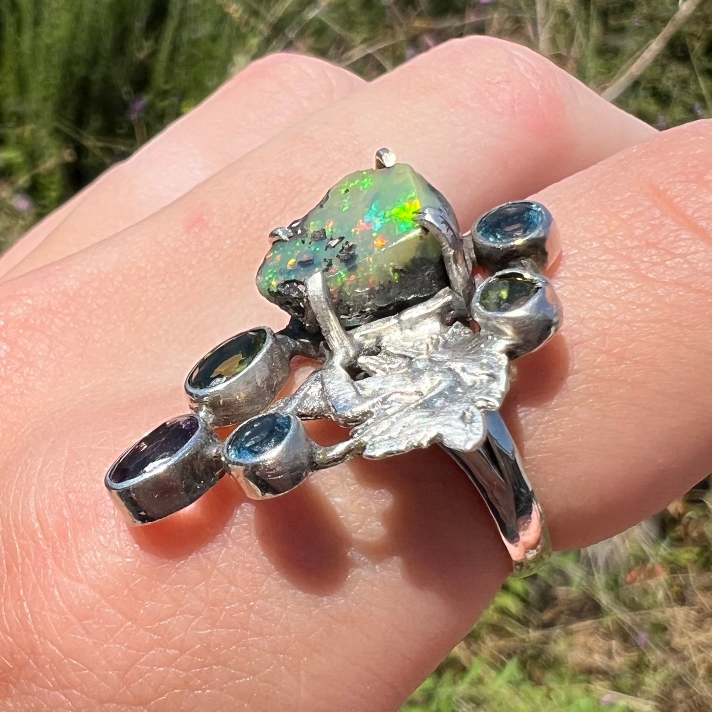A silver men's ring set with rough Virgin Valley opal and gems with a female figure design shown at an angle in sunlight.