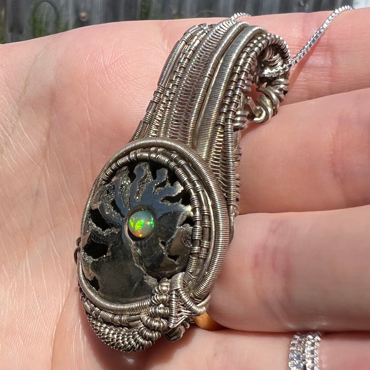 Man's wire wrapped ammonite pendant with Ethiopian opal accent that shows green flash in sunlight, shown held in hand from angle.